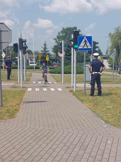 Uczeń przejeżdża rowerem chodnikiem, w tle widać zielone światło i dwóch policjantów obserwujących ucznia zdającego egzamin na kartę rowerową.