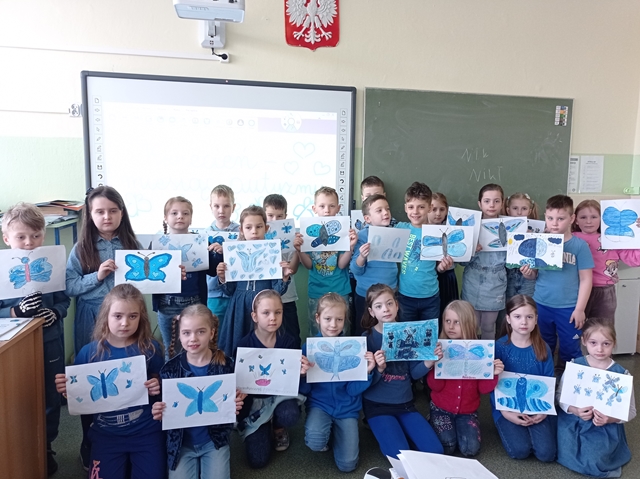 Grupa uczniów ubranych na niebiesko trzyma w rękach rysunki niebieskich motyli.