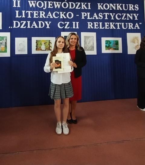 Uczennica w towarzystwie swojej nauczycielki prezentuje dyplom i nagrodę zdobytą w II Wojewódzkim Konkursie Literacko- Plastycznym „Dziady cz. II Relektura”. W tle wystawa prac plastycznych nagrodzonych w tym konkursie.