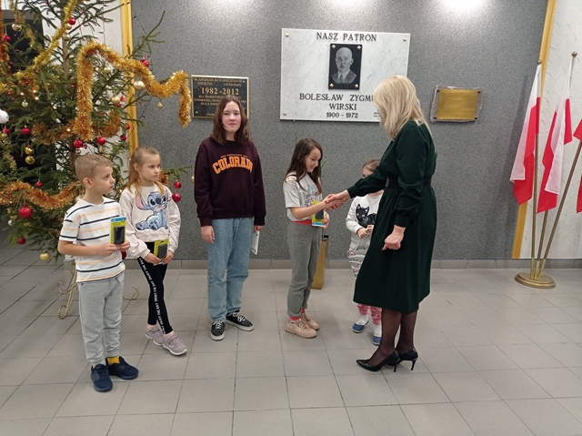 Zdjęcie przedstawia uczniów odbierających nagrody  od Pani Dyrektor za udział w konkursie na najpiękniejsze stroiki bożonarodzeniowe. W tle widać choinkę ubraną w złote i czerwone bombki oraz łańcuchy.