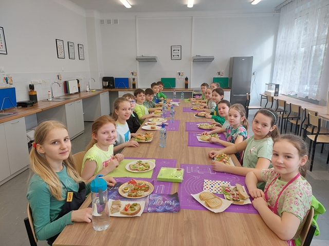 Zdjęcie przedstawia uczniów  w pracowni kulinarnej, siedzących przy stolikach wraz z przygotowanymi  przez siebie kanapkami.
