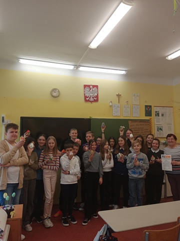 Zdjęcie przedstawia grupę uczniów w klasie wraz z nauczycielem. Uczniowie trzymają w ręku jajka wielkanocne wykonane podczas warsztatów artystycznych.