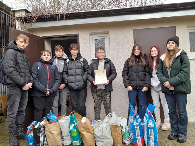 Zdjęcie przedstawia uczniów przekazujących dary dla zwierząt przebywających w schronisku. Młodzież pozuje do ujęcia z dyplomem i paczkami karmy. 