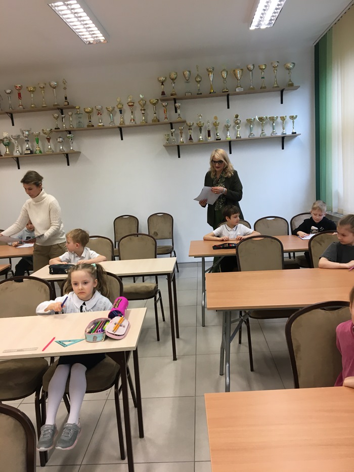 Uczniowie klas I–III siedzą przy stolikach, przygotowani do rozpoczęcia konkursu matematycznego. Pani Dyrektor wraz z Panią Wicedyrektor przechodzą między dziećmi, rozdając im arkusze z zadaniami.