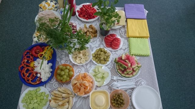 Zdjęcie przedstawia stół zastawiony różnymi owocami i warzywami. Wszystkie produkty są pokrojone, leżą na białych talerzykach, a wśród nich m.in. pomidory, mandarynki czy banany. Stół przystrojony jest w różne zielone zioła i kolorowe serwetki.
